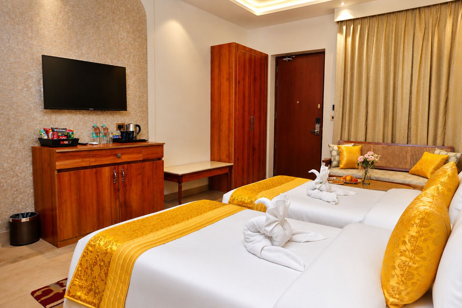 Ein Hotelzimmer mit Flachbildfernseher, Holzschrank, Schlafsofa mit gelb-weißer Bettwäsche und einem kleinen Tisch mit Blumenvase.