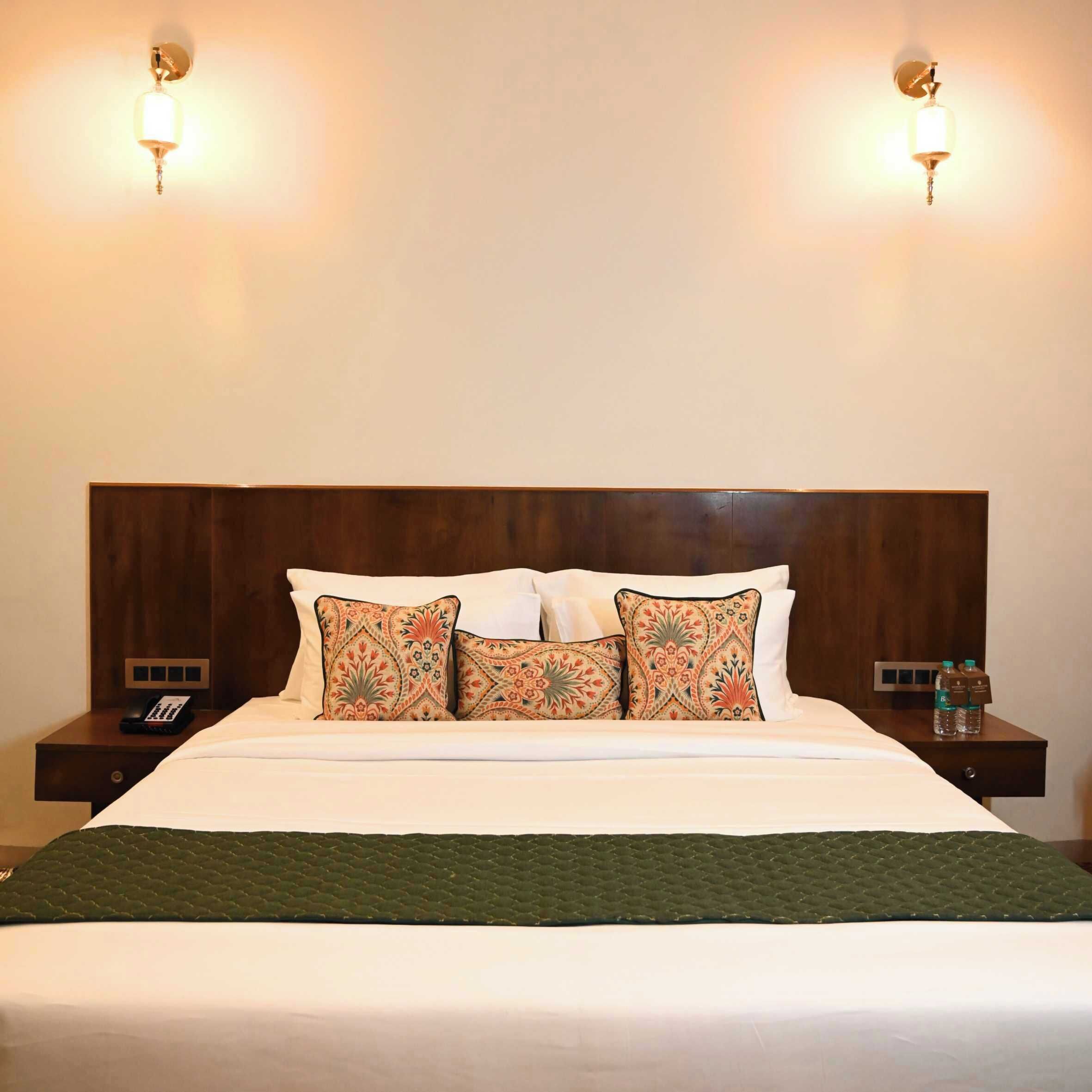 A neatly made bed with patterned pillows, a wooden headboard, bedside tables with a phone and water bottles, and wall-mounted lights.