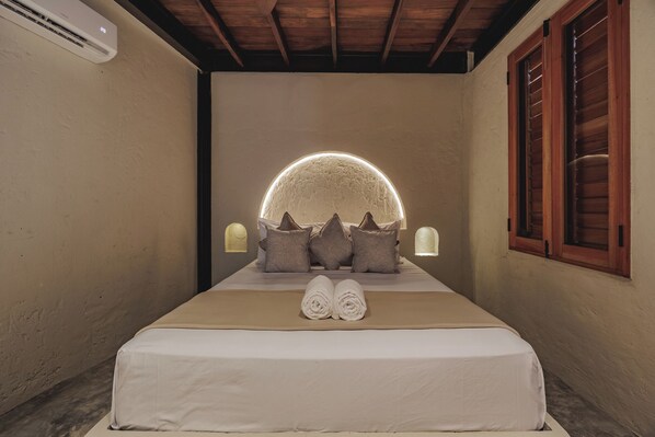 Une chambre à coucher avec un lit, des oreillers, une tête de lit dotée d’une lumière circulaire et une fenêtre avec des volets en bois.