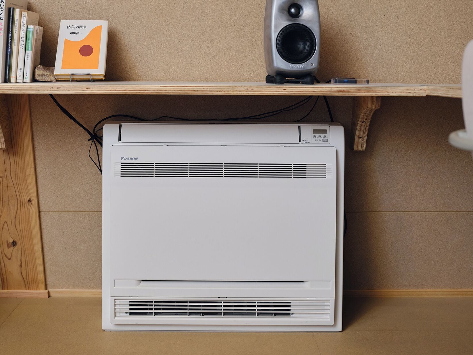 A room with a white air conditioner, a bookshelf with books and a book with an orange cover, and a speaker on a shelf.