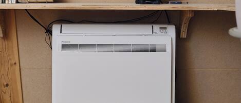 A room with a white air conditioner, a bookshelf with books and a book with an orange cover, and a speaker on a shelf.