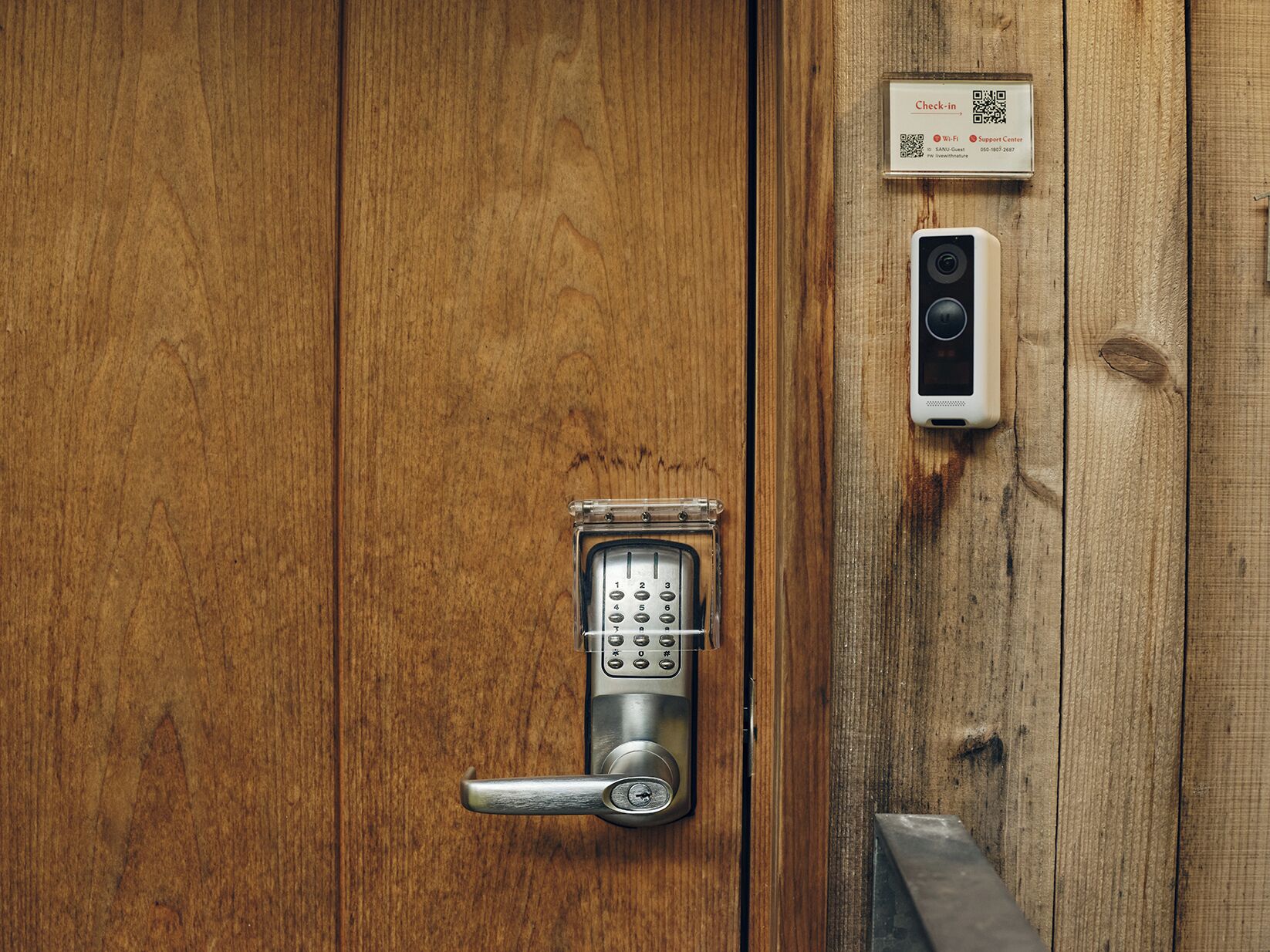 A wooden door with a numeric keypad lock, a QR code, and a small device on the wall.