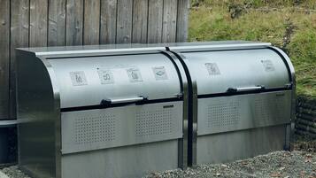 A metal recycling bin with labeled compartments for bottles, cans, and plastic bottles, placed outdoors with a wooden fence in the background.