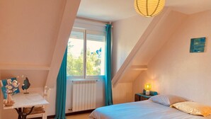 A bedroom with a bed, a table, a chair, a radiator, a window with curtains, and a hanging light fixture.