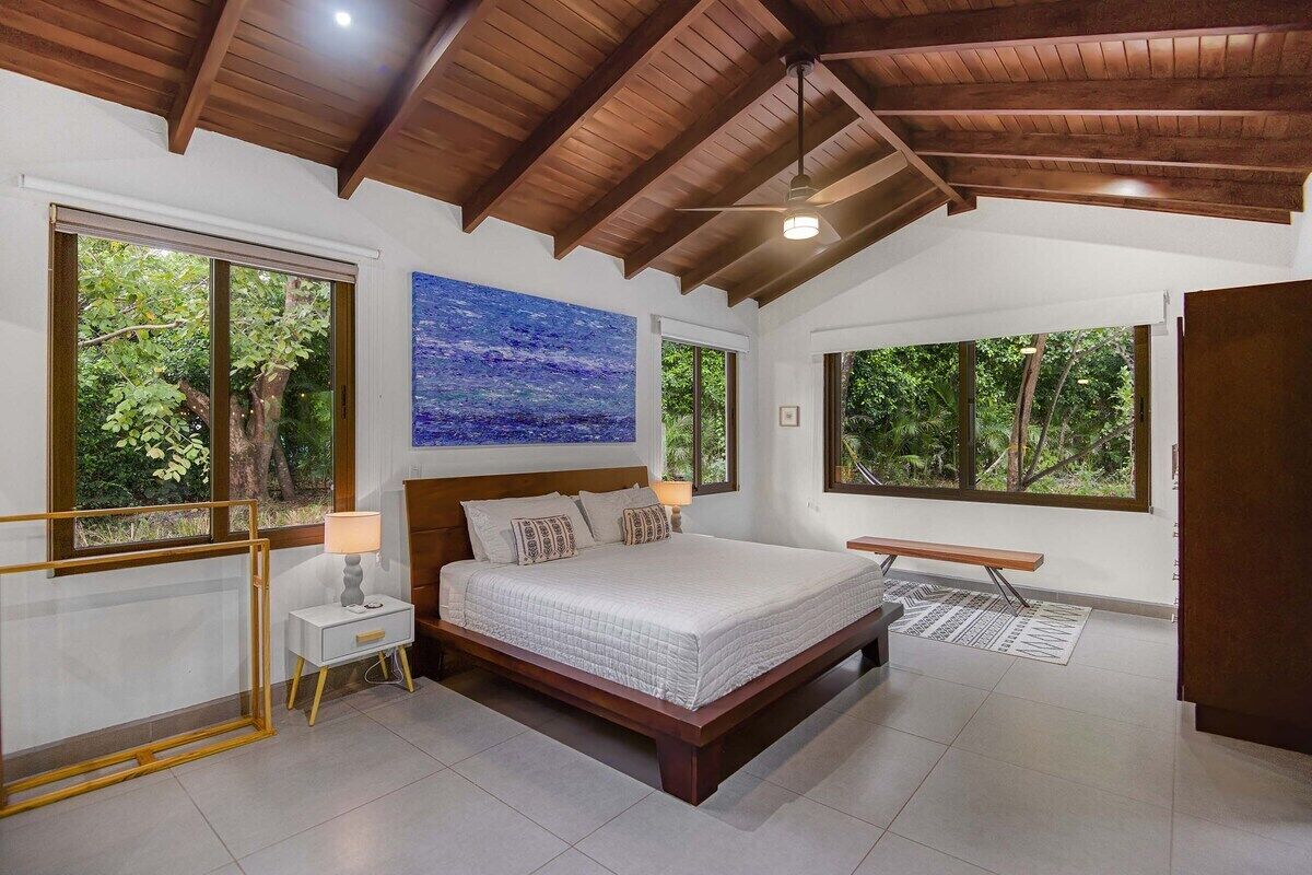 Un dormitorio con una cama grande, vigas de madera en el techo y ventanas amplias con vistas al verdor.