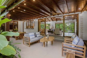 A spacious living area with a wooden ceiling, a sofa set, a coffee table, and a view of the outdoors through large windows.