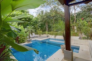 A swimming pool with a tiled edge, surrounded by lush greenery and a wooden pergola structure.