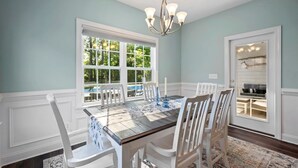 A dining room with a wooden table, white chairs, a chandelier, and a view of a backyard with a pool. - Pawleys Island Retreat with Private Pool (Pawleys Island)