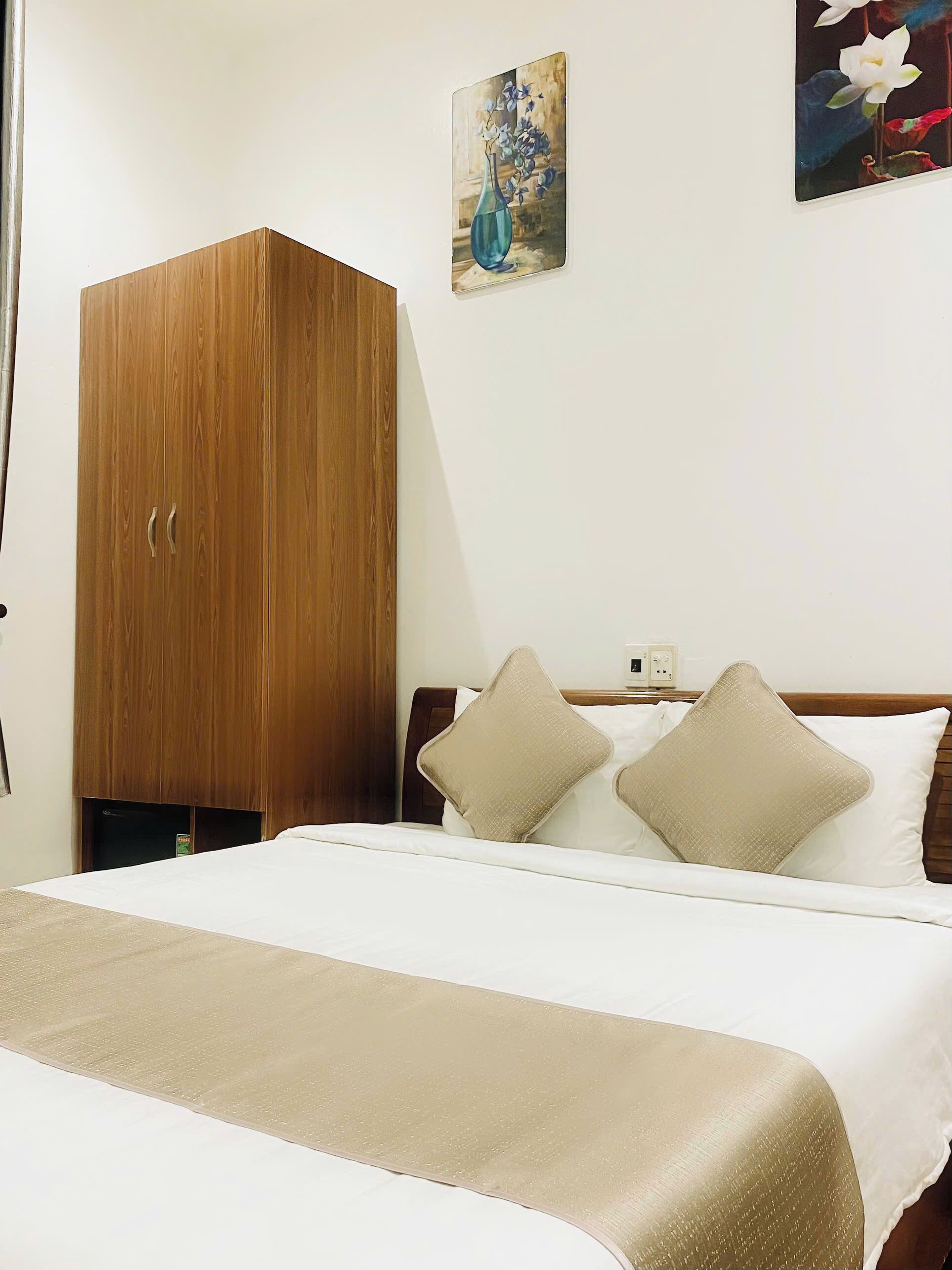 A hotel room with a wooden wardrobe, a bed with white bedding and beige pillows, and a painting of flowers on the wall.