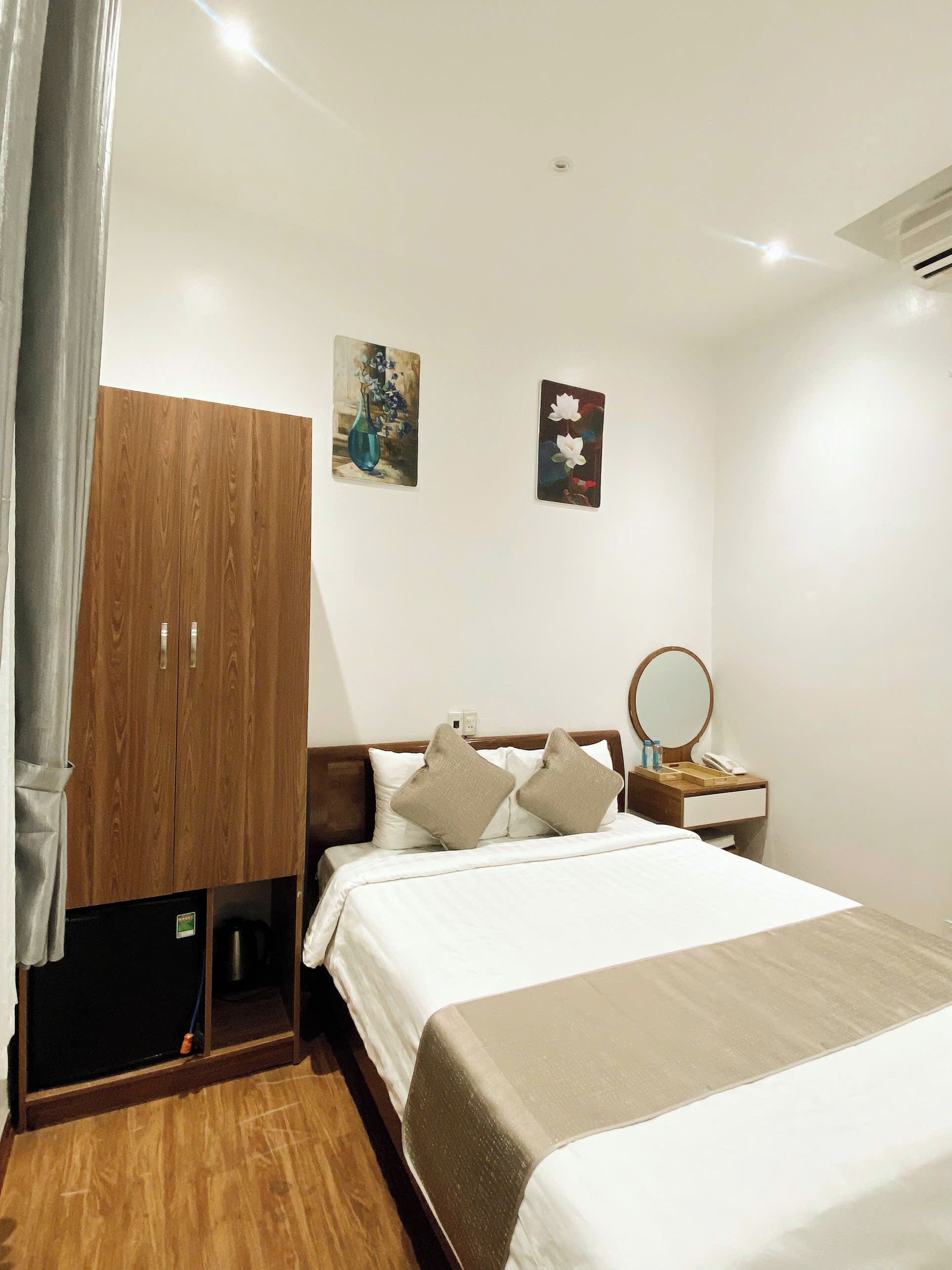 Quarto de hotel com guarda-roupa de madeira, cama com roupa de cama branca, mesa de cabeceira com espelho e duas pinturas penduradas na parede.