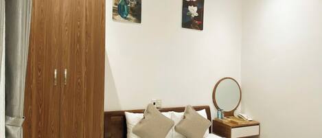 Quarto de hotel com guarda-roupa de madeira, cama com roupa de cama branca, mesa de cabeceira com espelho e duas pinturas penduradas na parede.