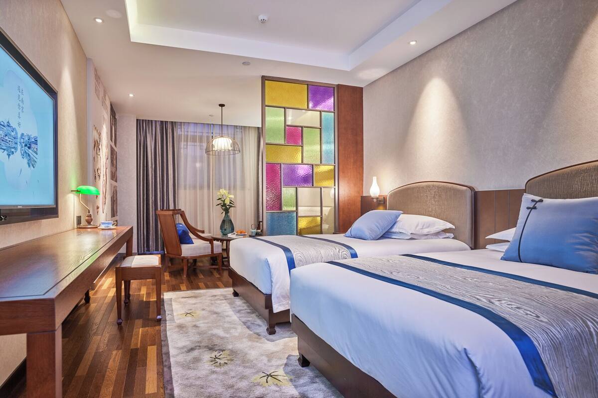 A hotel room with two beds, a desk, a chair, and a large window with curtains.