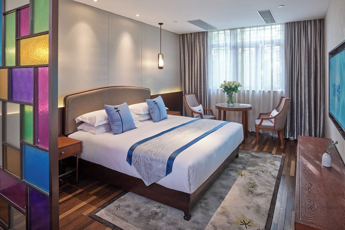 A hotel room with a large bed, two bedside tables, a small round table, two chairs, a TV, and a window with curtains.