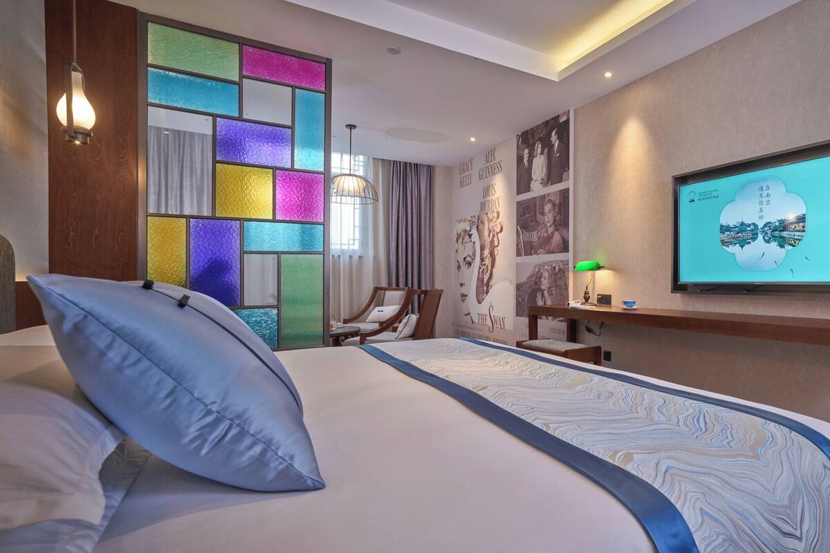 A hotel room with a bed, a desk, a chair, a TV, and a colorful wall panel.