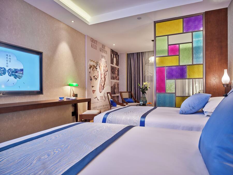 A hotel room with two beds, a desk with a lamp, a TV, and a colorful wall panel.