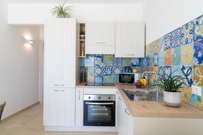 A compact kitchen with white cabinetry, a built-in oven, a microwave, and a tiled backsplash with a blue and yellow pattern. - GuestHost - Charming 40 sqm apartment, perfect for 4 people, located on the first floor of a building.The property is located inside the Borgo 81 Residence, in a quiet location and a few minutes drive from the center of Marzamemi and the main tourist des (Pachino)