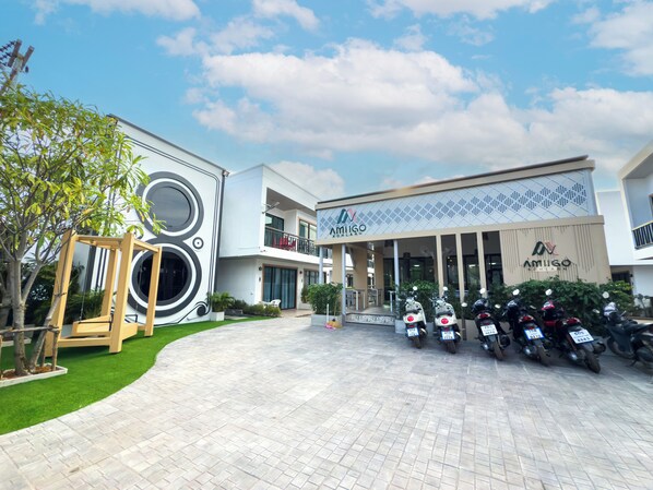 A modern building with a large circular design on the facade, a parking area with several scooters, and a small garden area with artificial grass and trees. - Amiigo Kohlarn (Koh Lan)