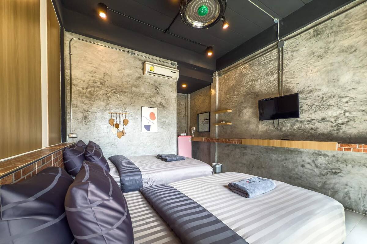 A modern hotel room with a large bed, a seating area with cushions, a mounted TV, and a decorative wall hanging.