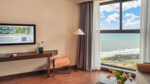 Habitación de hotel con escritorio de madera, una silla, vista al océano, ventana con cortinas y un mapa enmarcado en la pared.