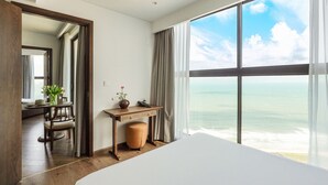 Habitación de hotel con un ventanal grande que ofrece vistas al océano, un escritorio de madera con un jarrón de flores, una silla y una cama.