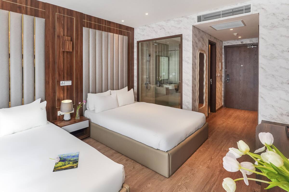 A modern hotel room with two beds, a nightstand, a lamp, and a bathroom visible through a glass partition.