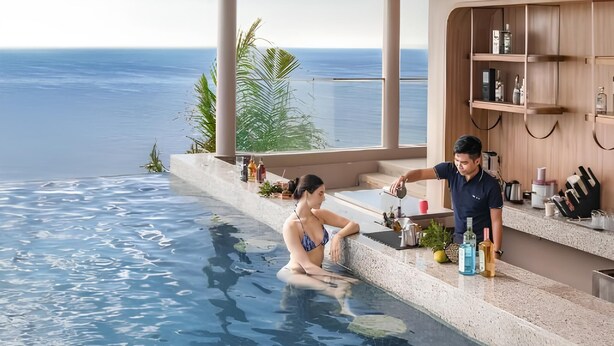 Zona de piscina moderna con vista al océano, terraza de madera y un bar con distintos artículos.