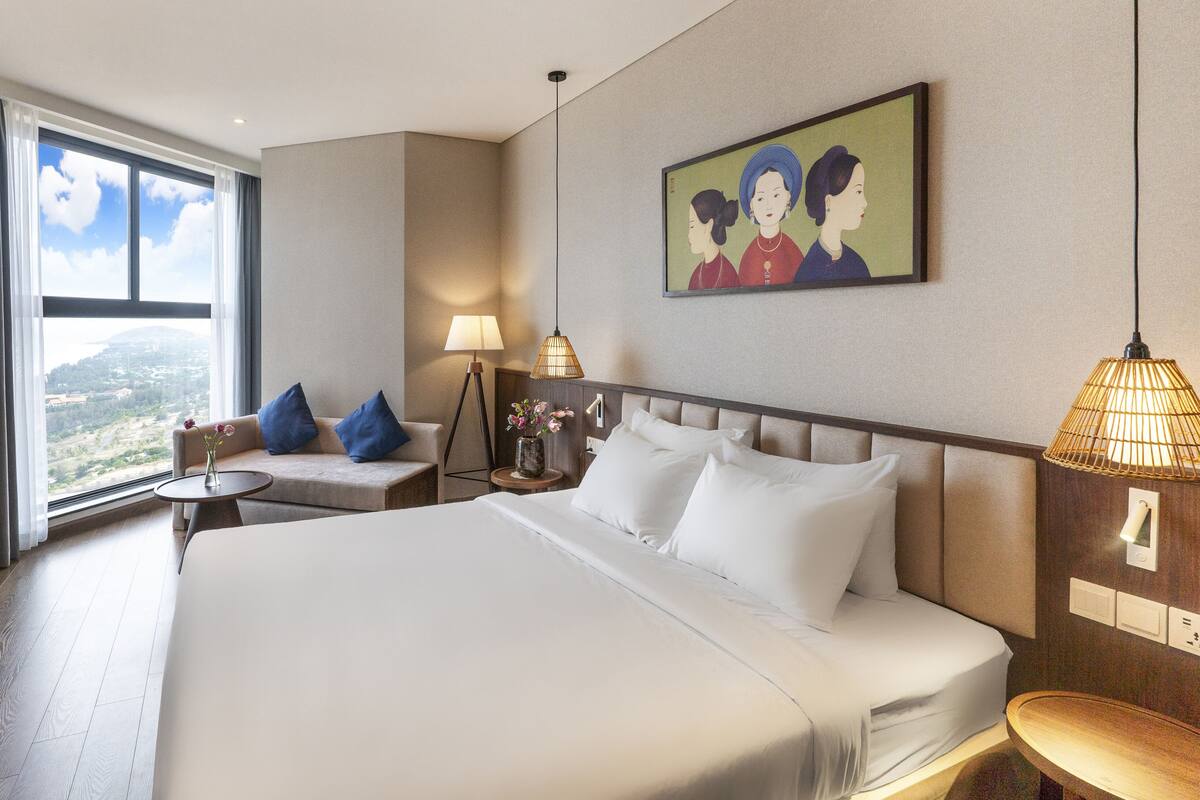 A hotel room with a large bed, a sofa, a small table with a vase of flowers, a lamp, and a painting on the wall.