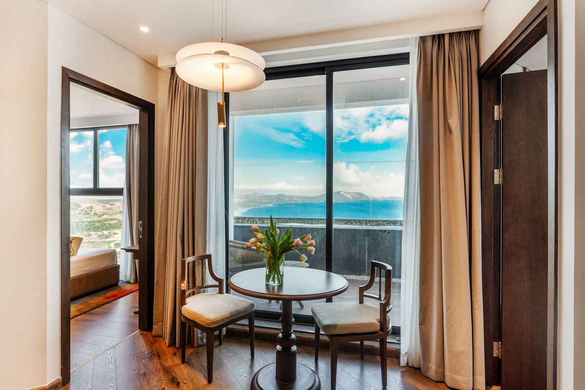 A room with a large window, a round table with chairs, a vase with flowers, and a view of the sea.