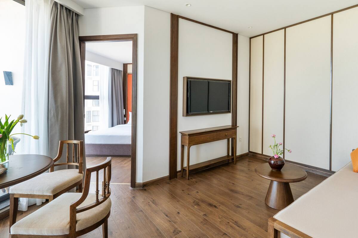 A hotel room with a wooden floor, a bed, a TV on a wooden stand, a small table with a vase, and a sofa with cushions.