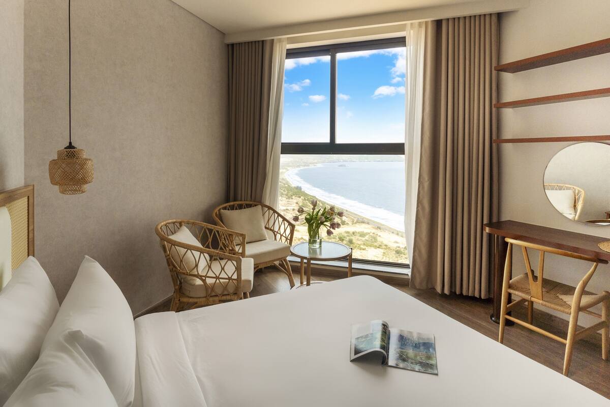 A hotel room with a bed, a desk, a chair, a round mirror, and a view of the beach.