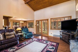 A spacious living room with a leather sofa, a blue armchair, a television, and a large window with blinds. - Conchas Cabin by Velo (Angel Fire)