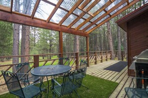 A covered outdoor deck with a table and chairs, surrounded by tall trees.
