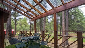 Un área de estar al aire libre cubierta, con una mesa y sillas, rodeada por una valla de madera y árboles altos.