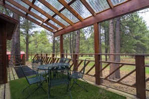 A covered outdoor seating area with a table and chairs, surrounded by a wooden fence and tall trees.