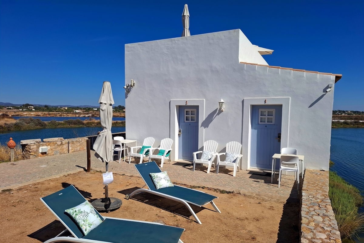 A small, white building with two doors and a patio area with lounge chairs and an umbrella.