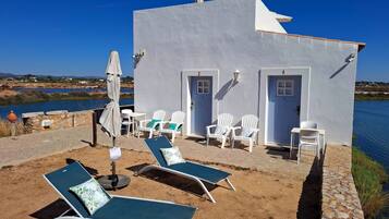 Un pequeño edificio blanco con dos puertas y un patio con sillas de descanso y una sombrilla.