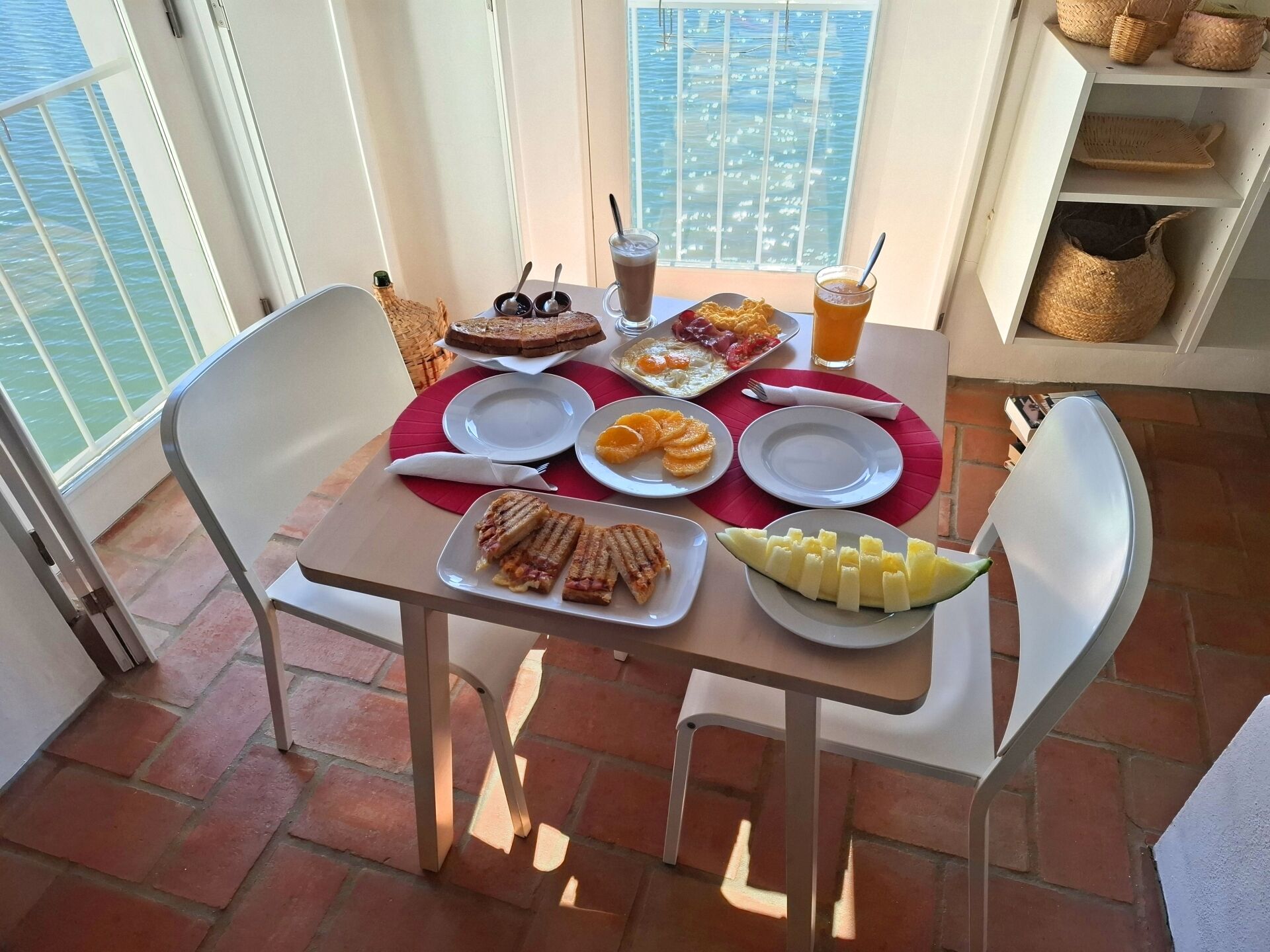 A balcony table with breakfast items, including toast, fruit, and beverages, overlooking a body of water.