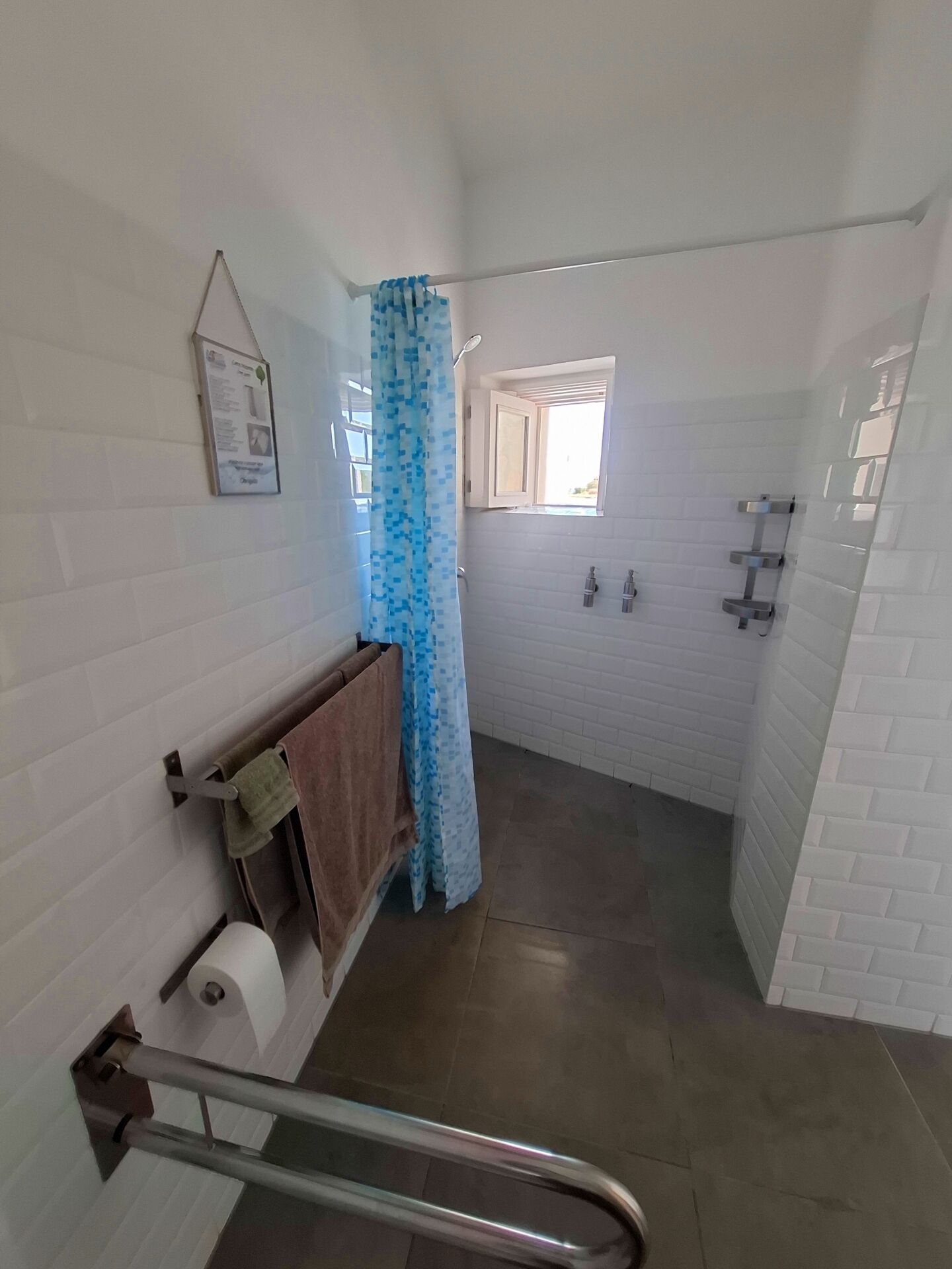 Un baño con paredes de azulejos blancos, una ducha con cortina azul, una ventana y un perchero para toallas.
