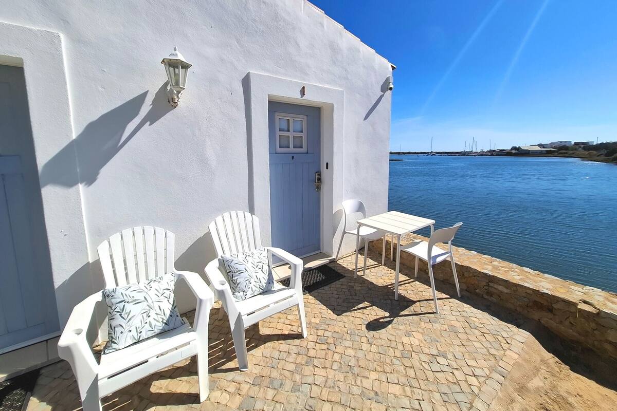 A white building with a door and windows, a paved patio with a table and chairs, and a view of the water.