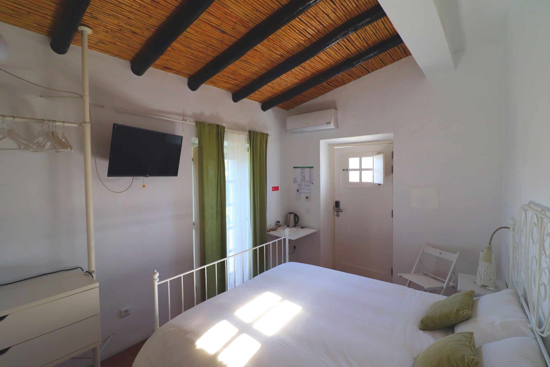 A bedroom with a bed, a TV mounted on the wall, a window with curtains, a door, and a radiator.