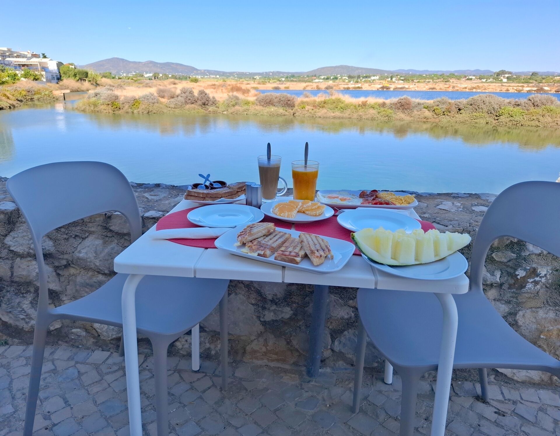 A table set with food and drinks by a lake, with chairs and a view of the water and hills in the background.