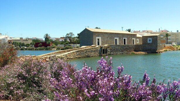 A stone building with a water body in the foreground and a clear sky. - Moinho das Marés (Olhao)