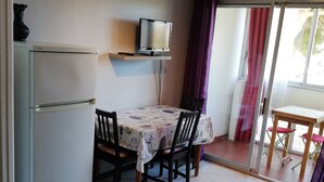 A compact room with a dining table, chairs, a television on a shelf, a refrigerator, and a sliding glass door leading to an outdoor area. - Studio for 2, fully equipped, loggia, park view, city center. (Balaruc-les-Bains)