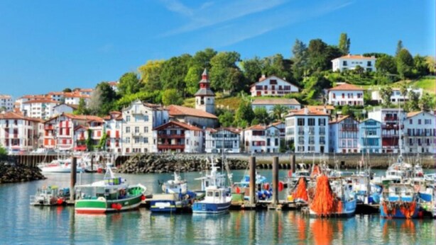 Un pueblo costero con edificios coloridos, un puerto deportivo con barcos y un faro al fondo.