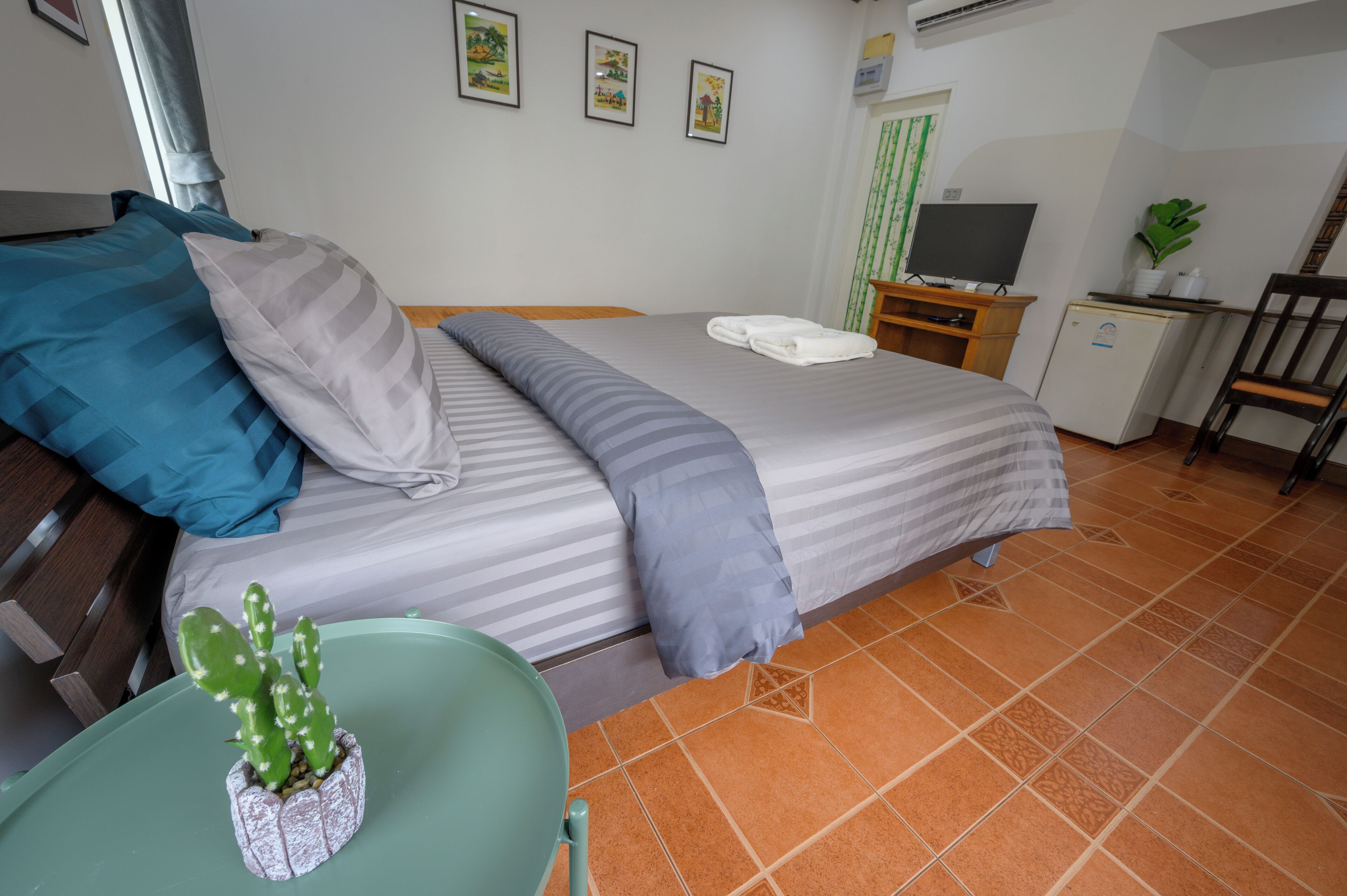 A neatly made bed with striped bedding, a small desk with a television, a green chair, and a cactus plant on the floor.
