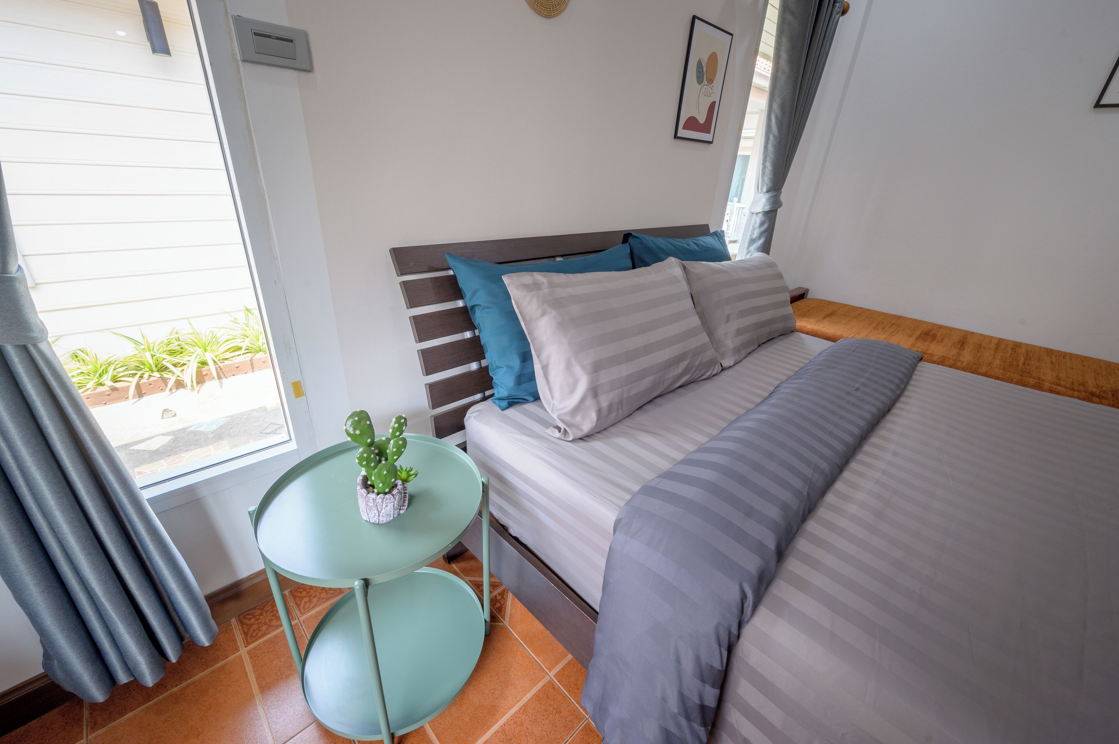 Een slaapkamer met een bed, een klein tafeltje met een cactus en een raam met gordijnen.