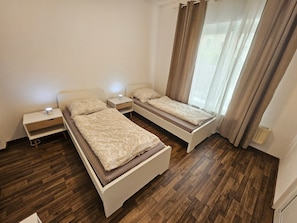 Una habitación con dos camas individuales, una mesita de noche con lámpara, un radiador y una ventana con cortinas.