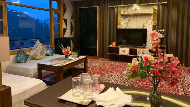 A hotel room with a large TV, a sofa, a dining table set with glasses and a vase of flowers, and a view of the mountains through the window.