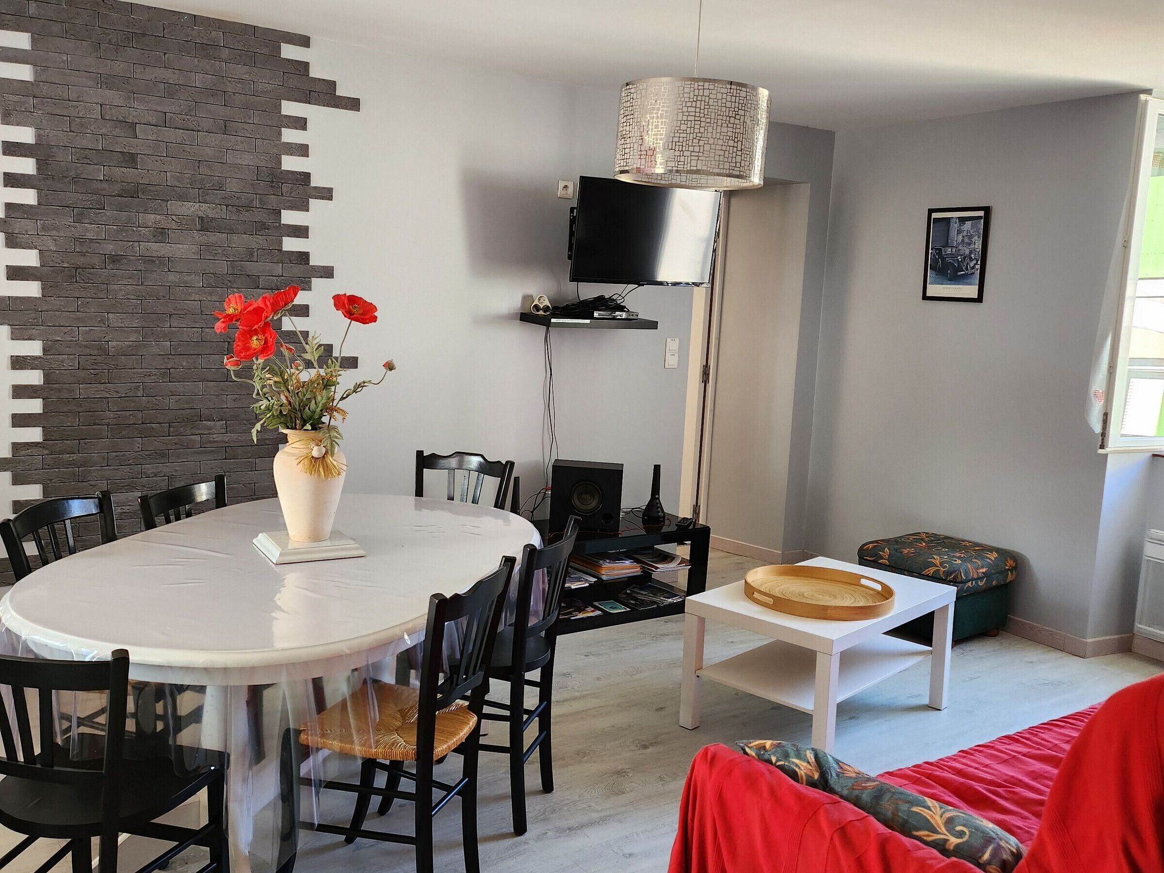 A modern dining area with a white round table, black chairs, a vase with red flowers, a brick-patterned wall, a black TV on a stand, and a small wooden table with a bowl.