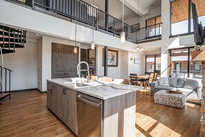 A modern kitchen with a central island, a dining area with a table and chairs, a staircase leading to an upper level, and a living area with a sofa and ottoman.